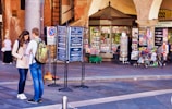 A man and a woman stand together on an urban street, possibly glancing at something in her hand. They are near a newsstand displaying numerous colorful magazines and newspapers. Bicycles are parked nearby, and a woman walks in the background under a building with brick arches.