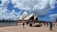 The image captures a view of the Sydney Opera House on a partially cloudy day. The iconic architecture with white, sail-like structures is prominently displayed against a vibrant blue sky filled with fluffy clouds. In the foreground, people are walking and enjoying the area, with a waterfront and cityscape visible in the background.