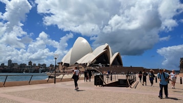 The image captures a view of the Sydney Opera House on a partially cloudy day. The iconic architecture with white, sail-like structures is prominently displayed against a vibrant blue sky filled with fluffy clouds. In the foreground, people are walking and enjoying the area, with a waterfront and cityscape visible in the background.
