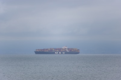 A cargo ship loaded with containers sailing across a calm ocean under a clear sky.