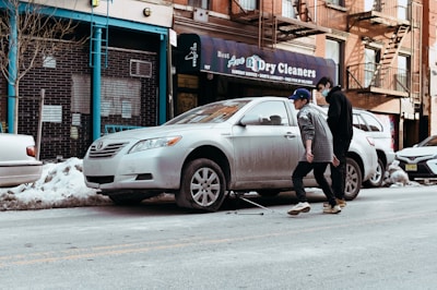Two people are walking on a city street beside a parked silver sedan. The car is missing a wheel and is supported by a jack, suggesting a tire change or repair. A storefront with a sign for a dry cleaning business is visible in the background, surrounded by snow piled along the sidewalk and curb. The setting appears urban with brick buildings and fire escapes.