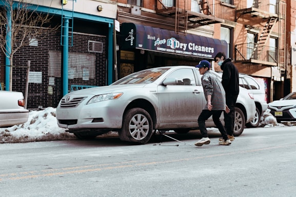 Two people are walking on a city street beside a parked silver sedan. The car is missing a wheel and is supported by a jack, suggesting a tire change or repair. A storefront with a sign for a dry cleaning business is visible in the background, surrounded by snow piled along the sidewalk and curb. The setting appears urban with brick buildings and fire escapes.