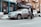 Two people are walking on a city street beside a parked silver sedan. The car is missing a wheel and is supported by a jack, suggesting a tire change or repair. A storefront with a sign for a dry cleaning business is visible in the background, surrounded by snow piled along the sidewalk and curb. The setting appears urban with brick buildings and fire escapes.