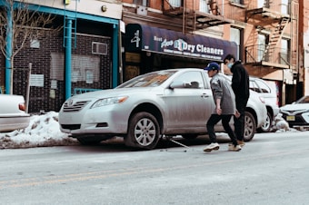 Two people are walking on a city street beside a parked silver sedan. The car is missing a wheel and is supported by a jack, suggesting a tire change or repair. A storefront with a sign for a dry cleaning business is visible in the background, surrounded by snow piled along the sidewalk and curb. The setting appears urban with brick buildings and fire escapes.