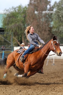 A woman is skillfully riding a horse at high speed in an outdoor setting. The horse is kicking up dust as it maneuvers sharply, showcasing impressive agility. In the background, blurred trees and a few spectators are visible.