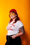 A person stands confidently against a bright orange background, wearing a red beanie, yellow-tinted sunglasses, a white t-shirt with red text, and black pants. They have long hair and a visible tattoo on their left arm. The person has an expression of self-assurance.