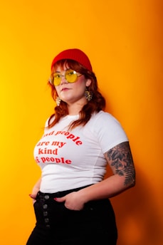 A person stands confidently against a bright orange background, wearing a red beanie, yellow-tinted sunglasses, a white t-shirt with red text, and black pants. They have long hair and a visible tattoo on their left arm. The person has an expression of self-assurance.