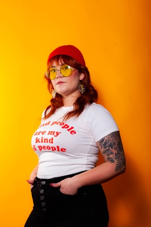 A person stands confidently against a bright orange background, wearing a red beanie, yellow-tinted sunglasses, a white t-shirt with red text, and black pants. They have long hair and a visible tattoo on their left arm. The person has an expression of self-assurance.