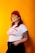 A person stands confidently against a bright orange background, wearing a red beanie, yellow-tinted sunglasses, a white t-shirt with red text, and black pants. They have long hair and a visible tattoo on their left arm. The person has an expression of self-assurance.