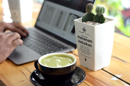 A person is typing on a laptop at a wooden table. In the foreground, a cup of green matcha latte is placed on a black saucer. Next to the cup, there is a white planter with small green cacti and a motivational quote in Indonesian.