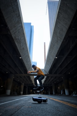 A person in a cap and yellow jacket is captured in mid-air above a one-wheeled electric skateboard on an empty urban street, framed by towering buildings and an underpass. The composition creates a dramatic perspective with the focus on motion and balance.
