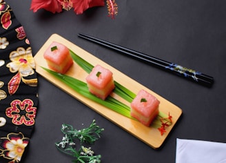 Three pink square-shaped edible items, possibly sushi or fruit, are arranged on a wooden board with green leaves underneath. Black chopsticks are placed to the side on a dark surface. A patterned fabric with floral designs and a red hibiscus flower are visible, adding a touch of elegance. Small decorative flowers enhance the composition.