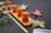 A bamboo board holds three pieces of rectangular, pink confectionery with small green toppings. Surrounding the setup are artificial, multicolored flowers and wooden chopsticks placed diagonally. In the background, a black cloth with intricate floral patterns contrasts with the dark grey surface.