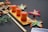 A bamboo board holds three pieces of rectangular, pink confectionery with small green toppings. Surrounding the setup are artificial, multicolored flowers and wooden chopsticks placed diagonally. In the background, a black cloth with intricate floral patterns contrasts with the dark grey surface.