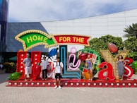 A group of people is standing in front of a large, festive display that reads 'Home for the Holidays.' The setup includes colorful, oversized letters and seasonal decorations like Christmas lights and ornaments. The background features a building with glass and a green hedge.