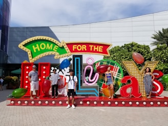 A group of people is standing in front of a large, festive display that reads 'Home for the Holidays.' The setup includes colorful, oversized letters and seasonal decorations like Christmas lights and ornaments. The background features a building with glass and a green hedge.