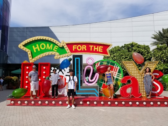 A group of people is standing in front of a large, festive display that reads 'Home for the Holidays.' The setup includes colorful, oversized letters and seasonal decorations like Christmas lights and ornaments. The background features a building with glass and a green hedge.