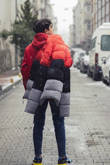 A person stands on a snowy urban street wearing a vibrant, multicolored puffer jacket and jeans. The street is lined with buildings and parked vehicles, with visible snowflakes adding a wintry atmosphere.