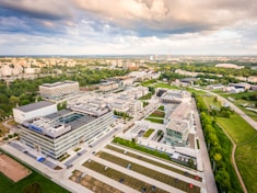 An aerial view of a large corporate or academic campus with multiple modern buildings, surrounded by lush greenery and open spaces. The sky is partly cloudy, with a warm sunlight glow washing over the scene. Roads, parking lots with parked cars, and walkways connect the structures. In the distance, a cityscape is visible on the horizon.