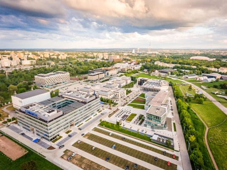 An aerial view of a large corporate or academic campus with multiple modern buildings, surrounded by lush greenery and open spaces. The sky is partly cloudy, with a warm sunlight glow washing over the scene. Roads, parking lots with parked cars, and walkways connect the structures. In the distance, a cityscape is visible on the horizon.