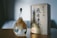 A uniquely shaped glass bottle with a cork sits on a wooden surface. The bottle contains a light amber liquid, and features a striking, detailed design of a mountain landscape at the base. Next to the bottle, there is a wooden box adorned with elegant Japanese calligraphy.