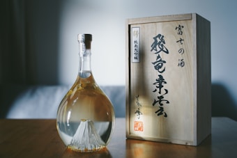 A uniquely shaped glass bottle with a cork sits on a wooden surface. The bottle contains a light amber liquid, and features a striking, detailed design of a mountain landscape at the base. Next to the bottle, there is a wooden box adorned with elegant Japanese calligraphy.