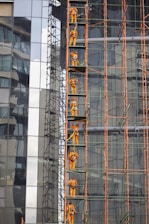 Construction workers in orange safety gear are climbing a scaffolding structure attached to a glass building. The scaffolding is composed of metal pipes, and the building features reflective glass panels, displaying urban architectural elements.