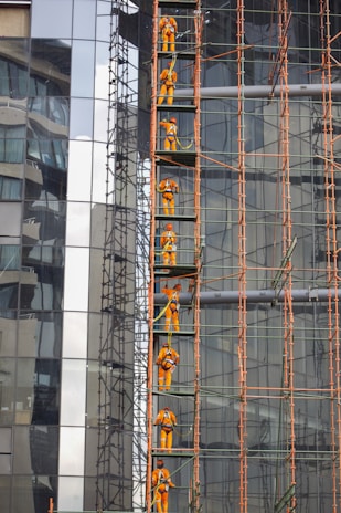 Construction workers in orange safety gear are climbing a scaffolding structure attached to a glass building. The scaffolding is composed of metal pipes, and the building features reflective glass panels, displaying urban architectural elements.