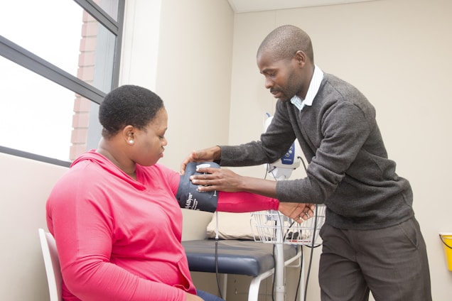A medical professional is checking the blood pressure of a seated woman in a clinical setting. The woman is wearing a pink shirt and has a blood pressure cuff on her arm. The room contains medical equipment and has a window allowing natural light.