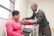 A medical professional is checking the blood pressure of a seated woman in a clinical setting. The woman is wearing a pink shirt and has a blood pressure cuff on her arm. The room contains medical equipment and has a window allowing natural light.