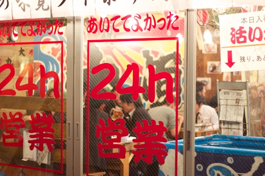 The image depicts the entrance of a restaurant with glass doors adorned with red Japanese characters indicating 24-hour service. Inside, several people are sitting at tables, engaging in conversation and dining. The interior features colorful wall art and appears lively and bustling.