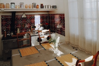 A cozy kitchen with vintage decor featuring a tiled backsplash in dark colors with intricate patterns. There are various ceramic jars and metal containers on the top shelf. A vase with white flowers is placed on a dining table, which is covered with a cloth and set with woven placemats. Wooden chairs surround the table, and soft, natural light filters through sheer curtains.
