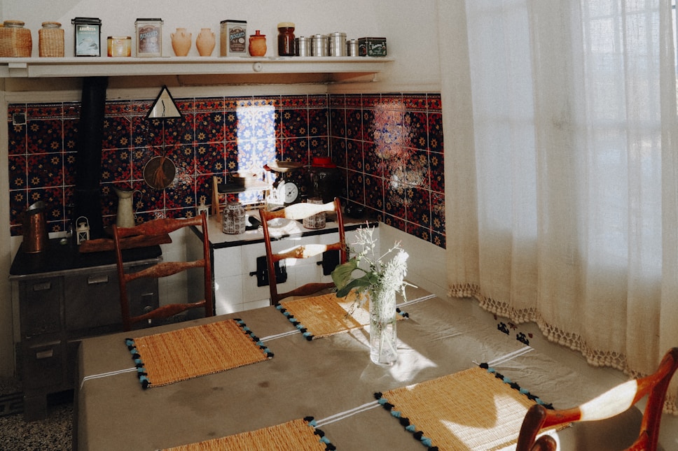 A cozy kitchen with vintage decor featuring a tiled backsplash in dark colors with intricate patterns. There are various ceramic jars and metal containers on the top shelf. A vase with white flowers is placed on a dining table, which is covered with a cloth and set with woven placemats. Wooden chairs surround the table, and soft, natural light filters through sheer curtains.