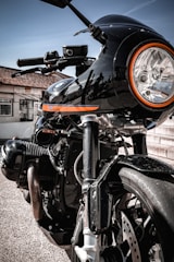 A close-up view of a black motorcycle with a prominent round headlight encircled by an orange ring. The bike's sleek and polished body features metallic accents and various mechanical components, with sunlight reflecting off the surface. The background includes a building facade with a tiled roof and a brick surface.