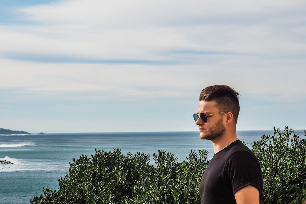 man in black crew neck t-shirt standing near green plants and body of water during