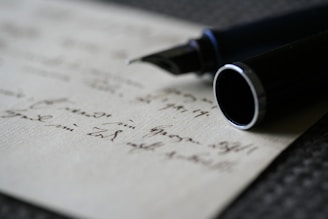 A close-up view of a fountain pen positioned atop a piece of paper with handwritten notes. The focus is on the nib of the pen, with the writing appearing slightly blurred in the background. The pen's dark body contrasts with the light color of the paper.