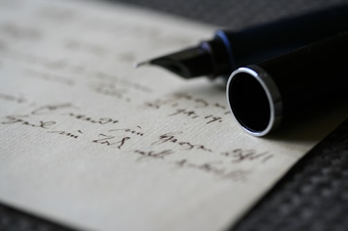 A close-up view of a fountain pen positioned atop a piece of paper with handwritten notes. The focus is on the nib of the pen, with the writing appearing slightly blurred in the background. The pen's dark body contrasts with the light color of the paper.