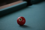A red billiard ball with the number 3 prominently displayed sits on a teal pool table. The background features the edge of the pool table with shadows creating a calm atmosphere.