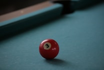 A red billiard ball with the number 3 prominently displayed sits on a teal pool table. The background features the edge of the pool table with shadows creating a calm atmosphere.