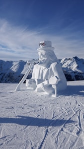 A large snow sculpture of a musician, complete with long hair, a beard, sunglasses, and a hat. The figure is playing a guitar. The background features snow-covered mountains under a clear blue sky.