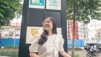 A young woman with glasses and a white ruffled blouse stands in front of a large sign with logos, smiling. The background includes urban elements such as a tree, buildings, and a street with vehicles.