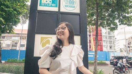 A young woman with glasses and a white ruffled blouse stands in front of a large sign with logos, smiling. The background includes urban elements such as a tree, buildings, and a street with vehicles.