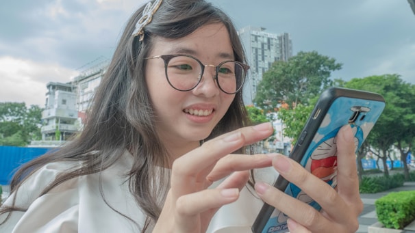 A young woman happily using her phone outdoors with a city skyline behind her.