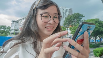 A young woman with glasses is smiling while using a smartphone. She has a decorative pin in her hair and wears a white blouse. The smartphone case features a cartoon character. The background shows an urban environment with buildings and trees.