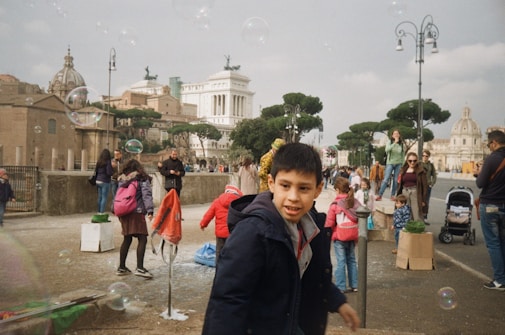 A bustling street with people engaging in various activities, bubbles floating in the air, and historic buildings in the background. Children are playing, and adults are walking and talking. A lively and joyful atmosphere is evident.