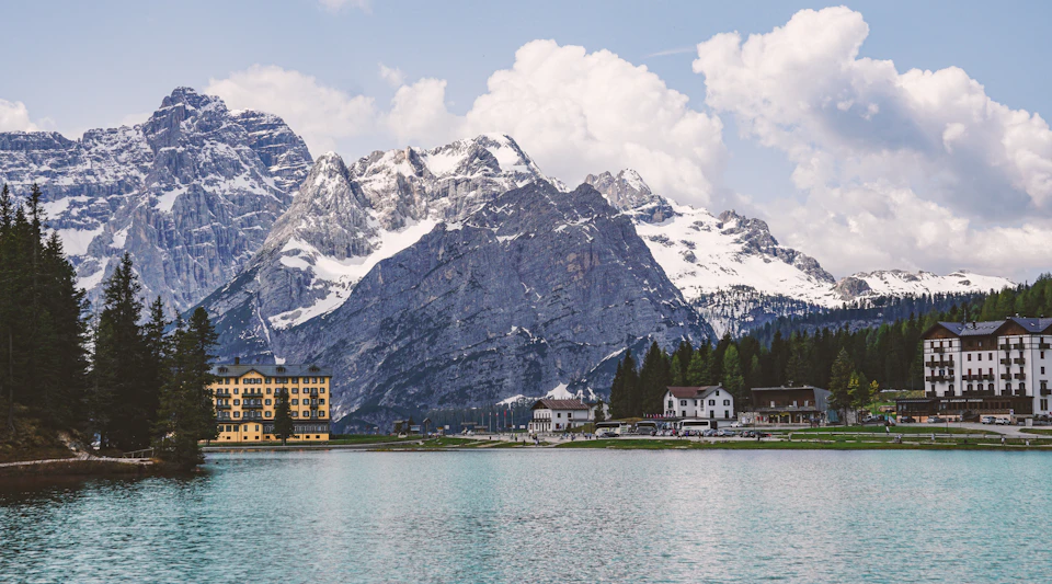 The image depicts a serene mountain landscape with a large body of water, possibly a lake or river, nestled between two snow-capped mountains. The mountains are covered in snow and appear to be part of a larger range, suggesting a high altitude location. In the foreground, there is a building that appears to be a hotel or resort, adding an element of human presence to the natural landscape. The sky above is clear blue with scattered clouds, creating a picturesque backdrop for the scene.
