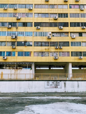 A multi-story building facade with a worn yellow exterior. Rows of windows, many with air conditioning units mounted alongside them. A sign in both English and Chinese directs towards a lift lobby. The walls appear aged with visible rust and stains.