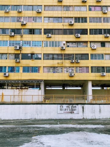 A multi-story building facade with a worn yellow exterior. Rows of windows, many with air conditioning units mounted alongside them. A sign in both English and Chinese directs towards a lift lobby. The walls appear aged with visible rust and stains.