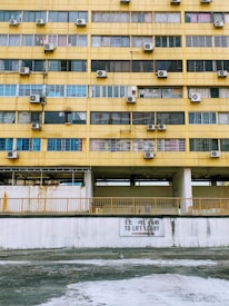 A multi-story building facade with a worn yellow exterior. Rows of windows, many with air conditioning units mounted alongside them. A sign in both English and Chinese directs towards a lift lobby. The walls appear aged with visible rust and stains.