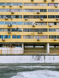 A multi-story building facade with a worn yellow exterior. Rows of windows, many with air conditioning units mounted alongside them. A sign in both English and Chinese directs towards a lift lobby. The walls appear aged with visible rust and stains.